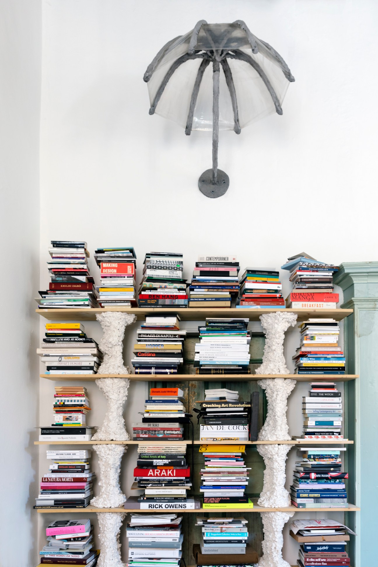 Transparent umbrella sculpture mounted on wall above wooden bookshelf with stacked books and decorative white supports