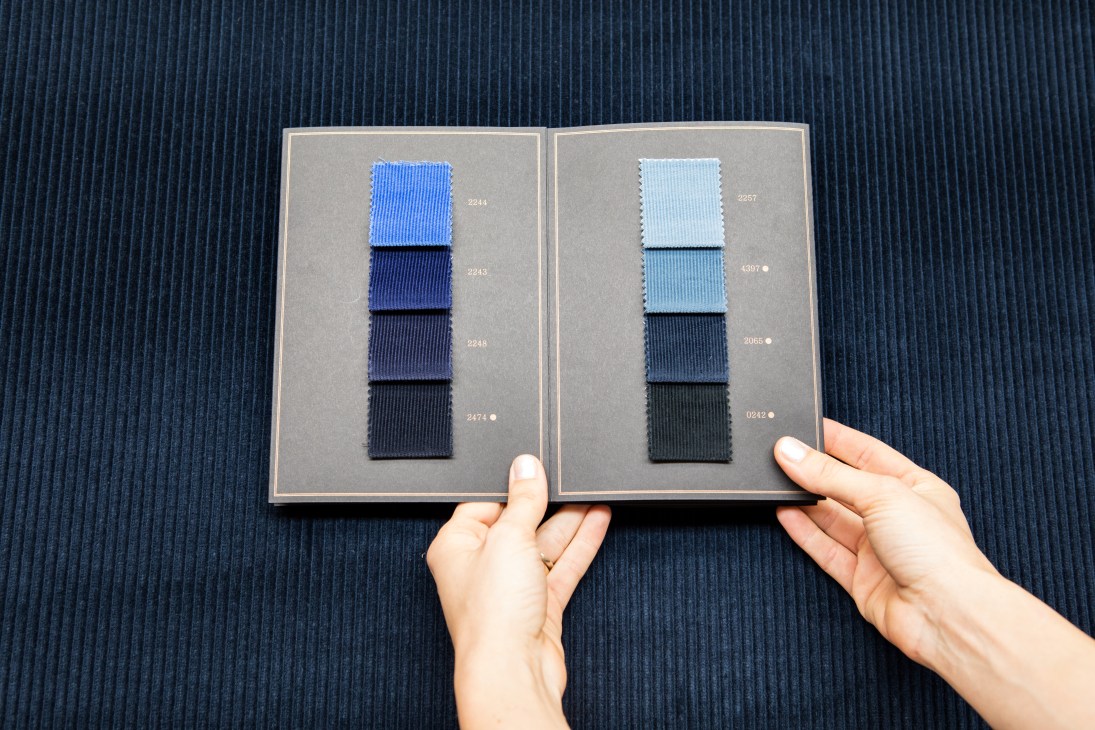 Hands holding open a fabric sample book displaying corduroy swatches in various shades of blue with numbered labels