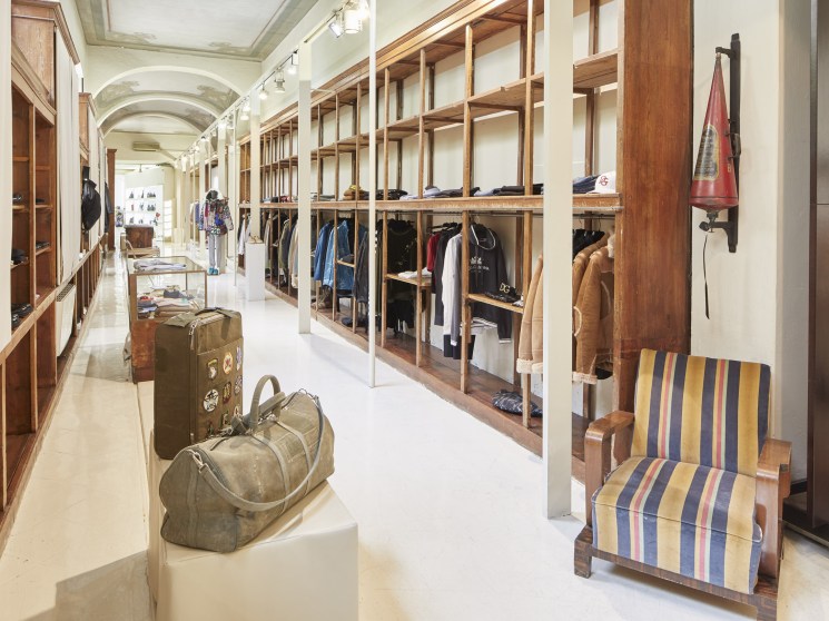 A long, elegant menswear boutique with warm wood shelving, hanging garments, vintage luggage displays, and a striped armchair.