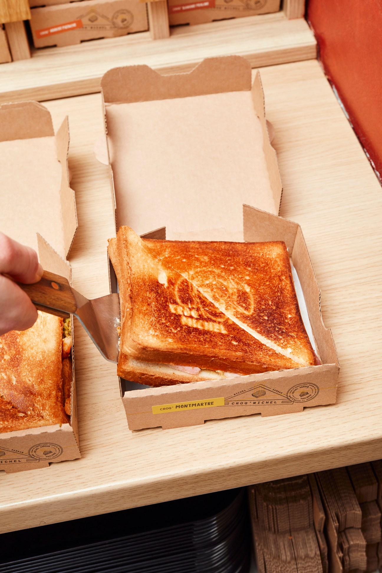 Golden grilled croque monsieur sandwiches in cardboard takeaway boxes being cut with a knife on wooden surface