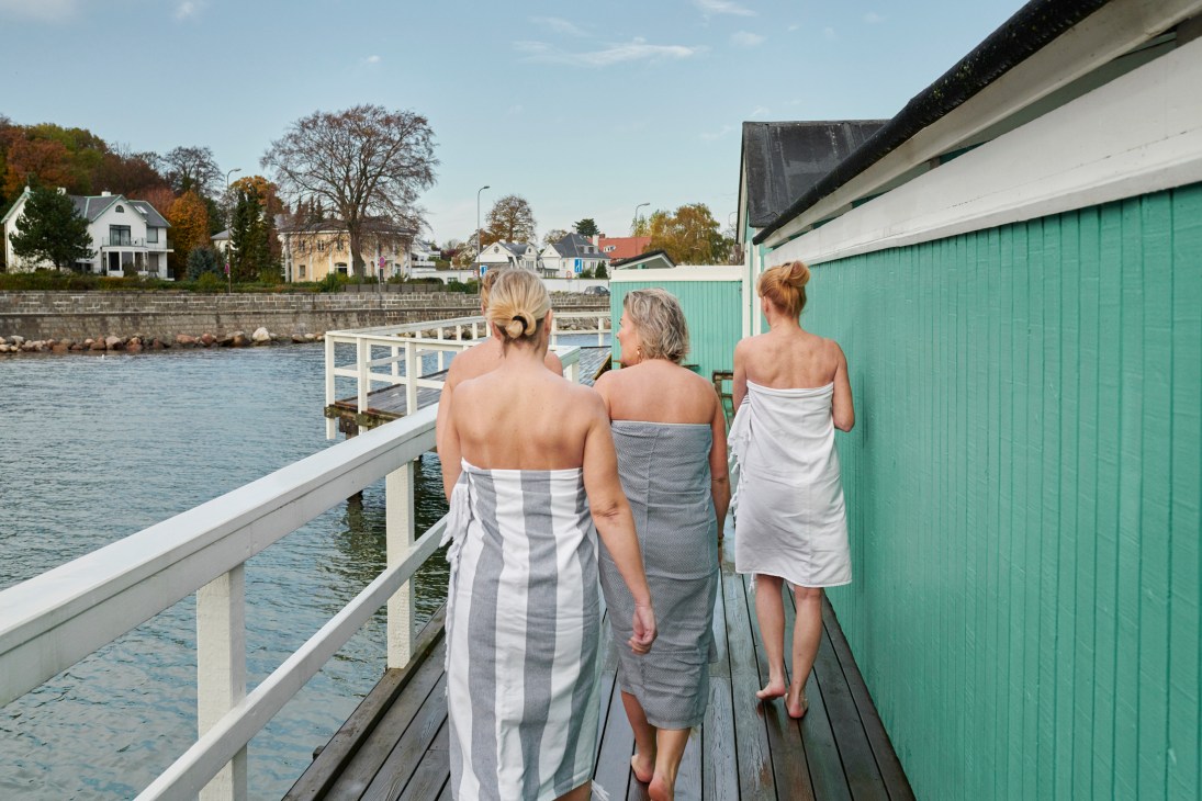 Three women wrapped in towels walk along a wooden dock toward the water at Charlottenlund Søbad winter-bathing club.