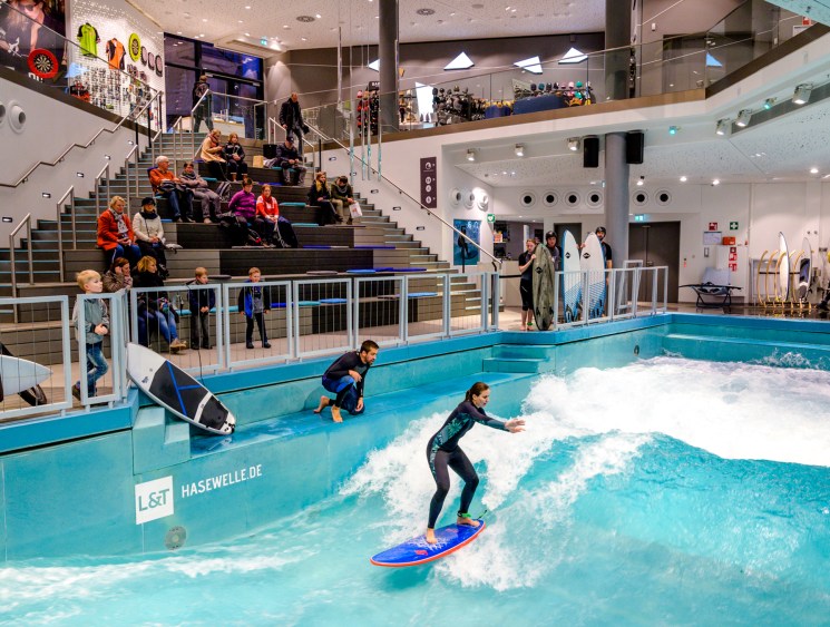 A surfer rides an artificial wave at an indoor surf facility while an instructor crouches beside the pool, with spectators watching from tiered seating.