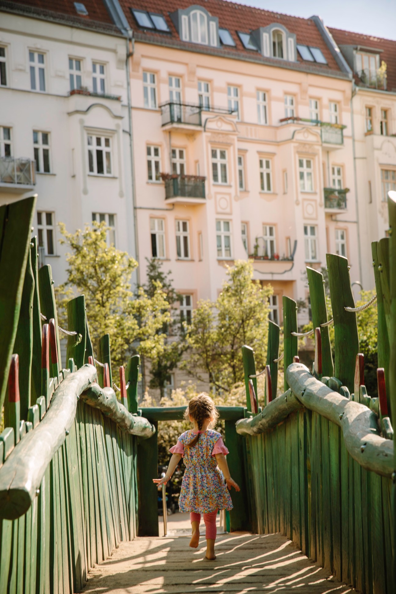 Child walking across a wooden rope bridge with green railings in a Berlin playground.