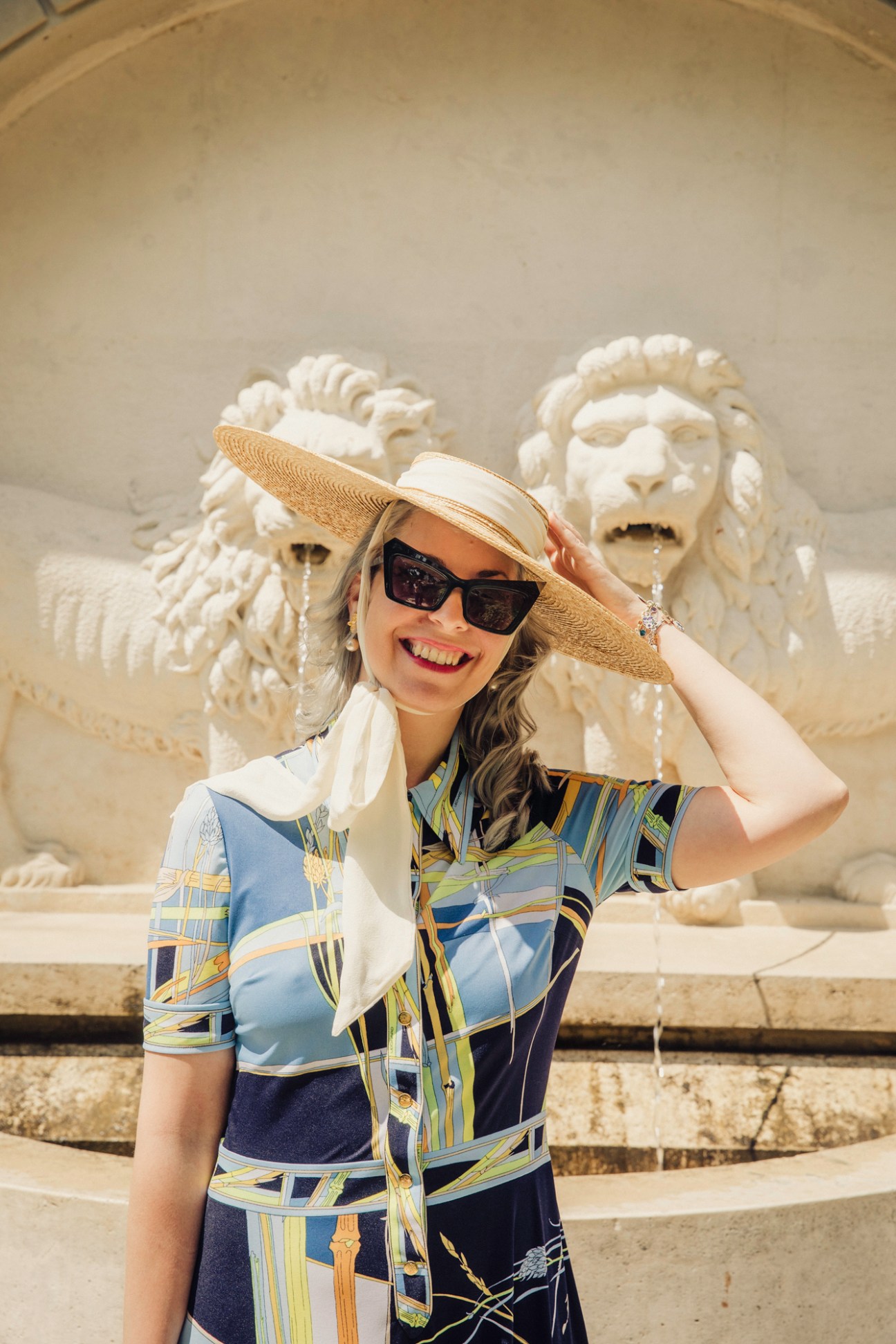 Woman in patterned dress and sun hat posing in front of ornate white stone fountain with sculptural figures
