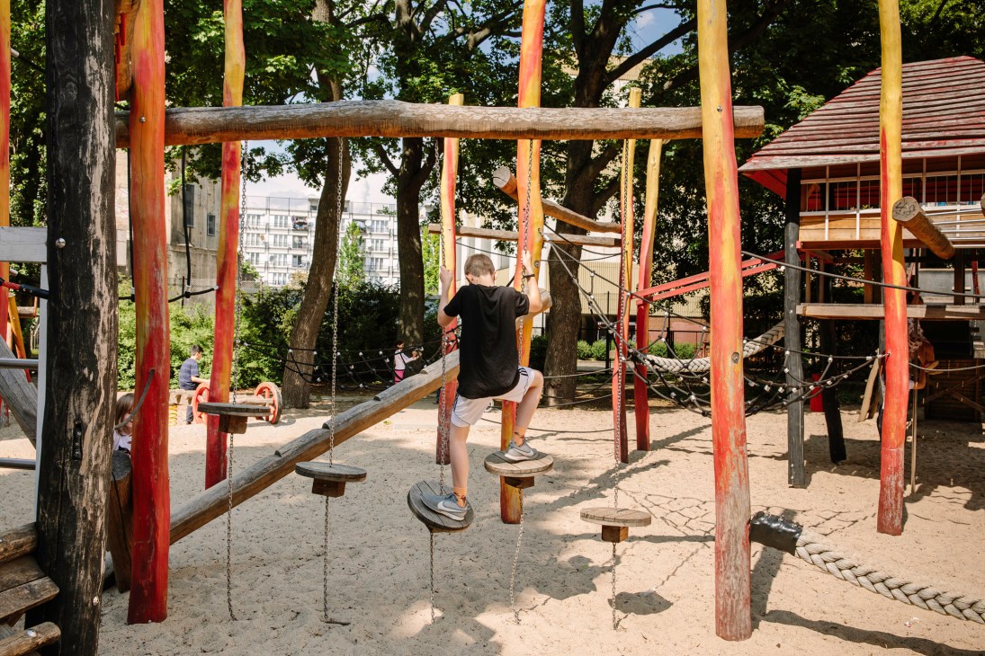 Child balancing on wooden steps suspended by chains in a playground with red posts and natural trees.