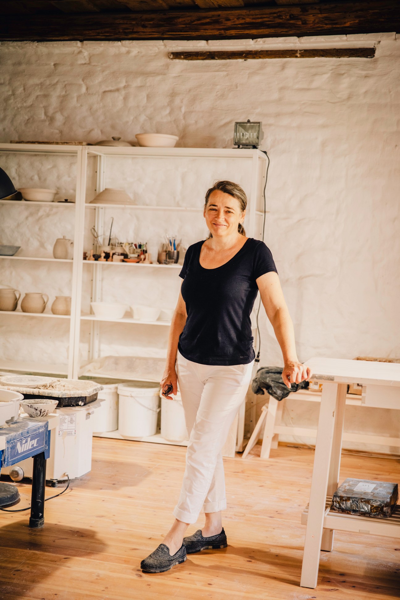 Potter Krisztina Szente standing in her ceramic workshop with pottery shelves and tools visible