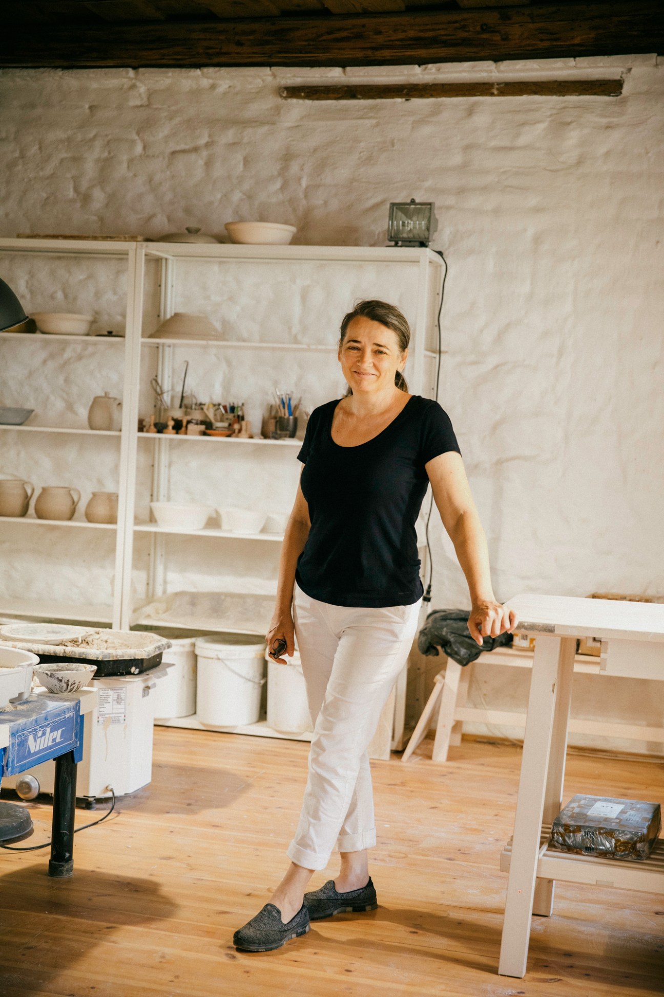 Ceramicist Krisztina Szente standing in her studio with white shelving and pottery supplies behind her.