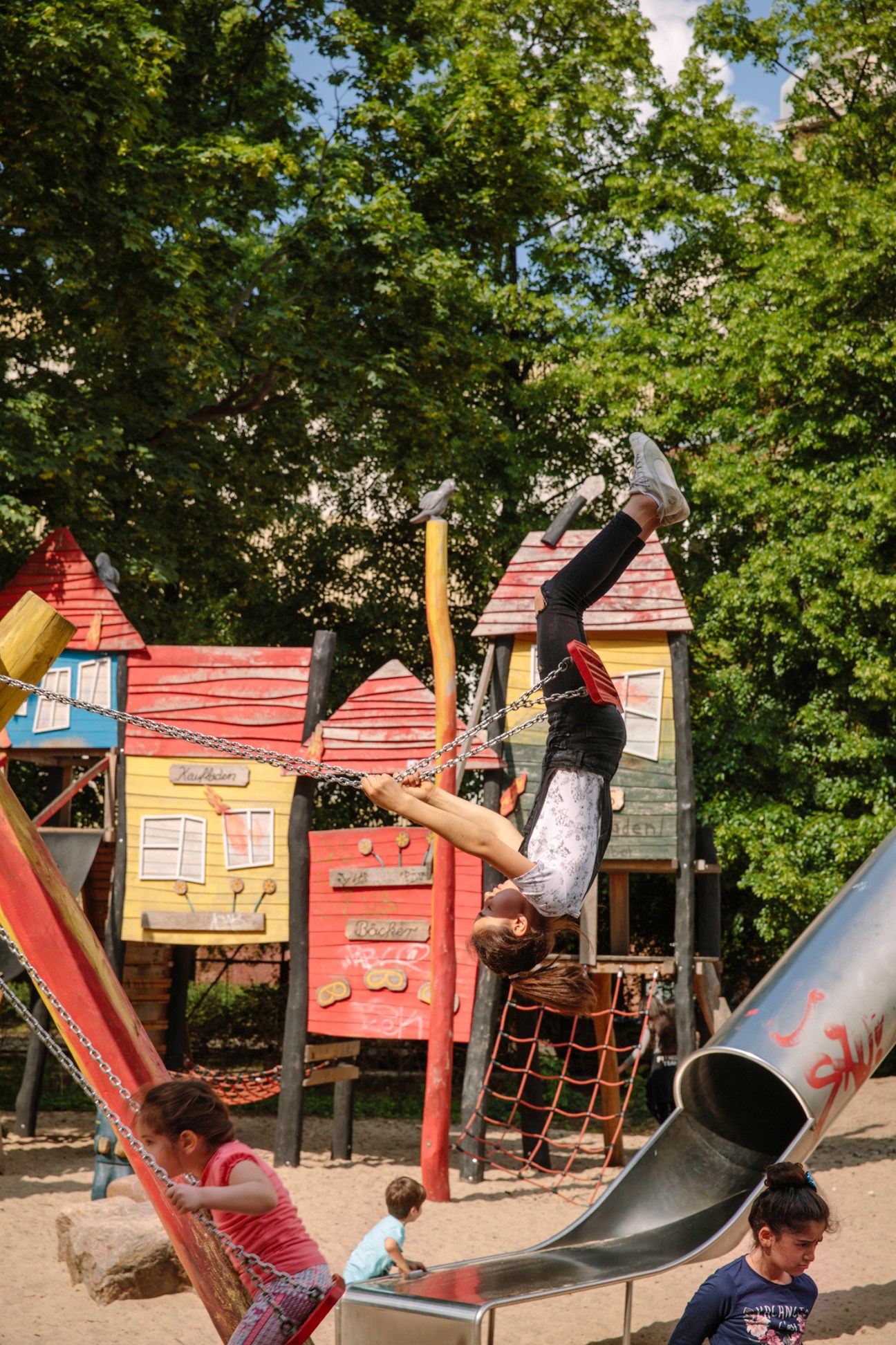Child hanging upside down from a rope swing at a colorful wooden playground with small houses and slide.