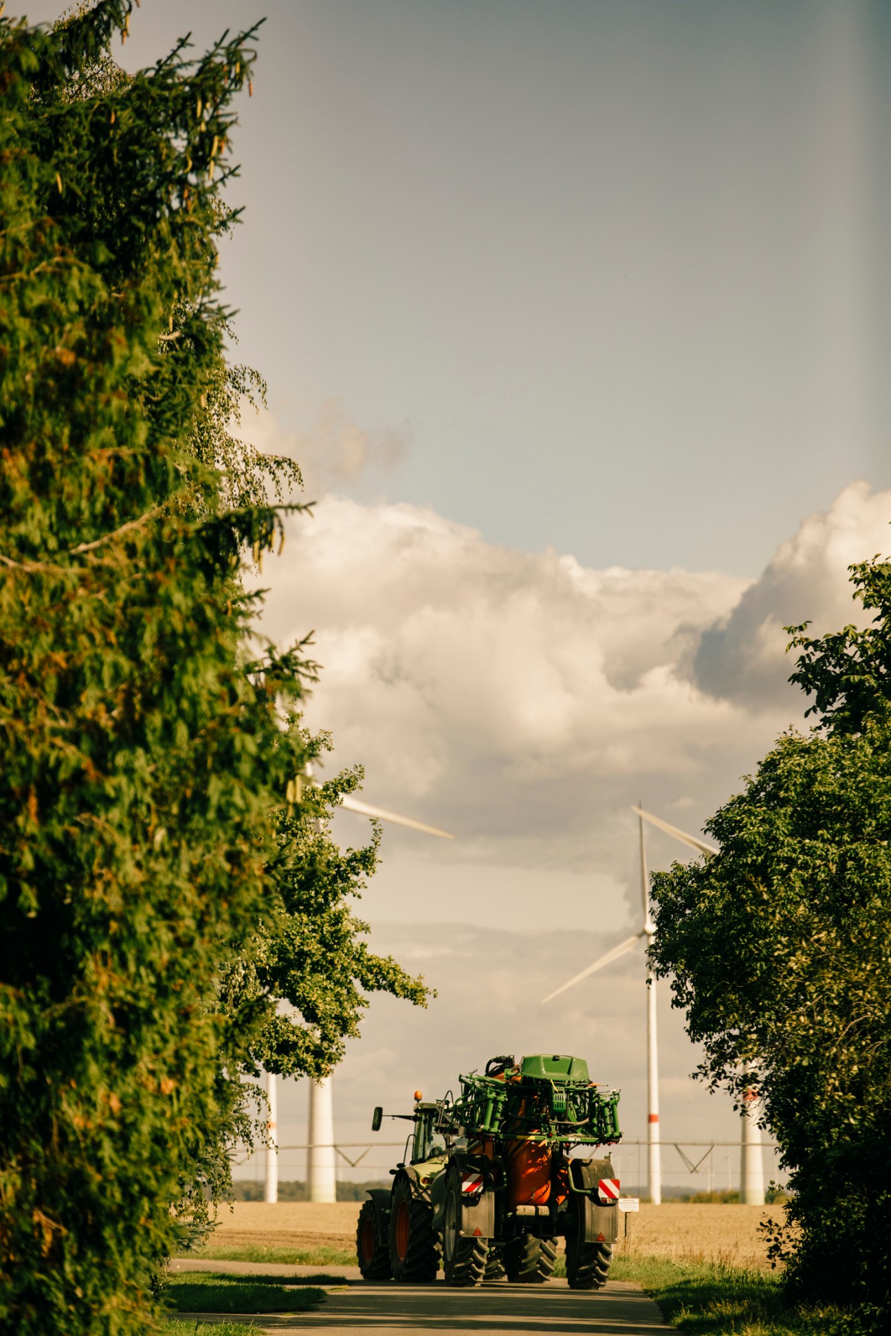 A green tractor drives on a rural road lined with vegetation, with wind turbines visible in the background across flat farmland.