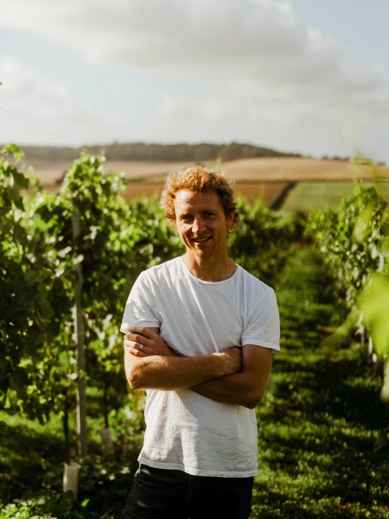 Daniel Ham stands smiling with arms crossed in front of lush green vineyard rows under cloudy sky