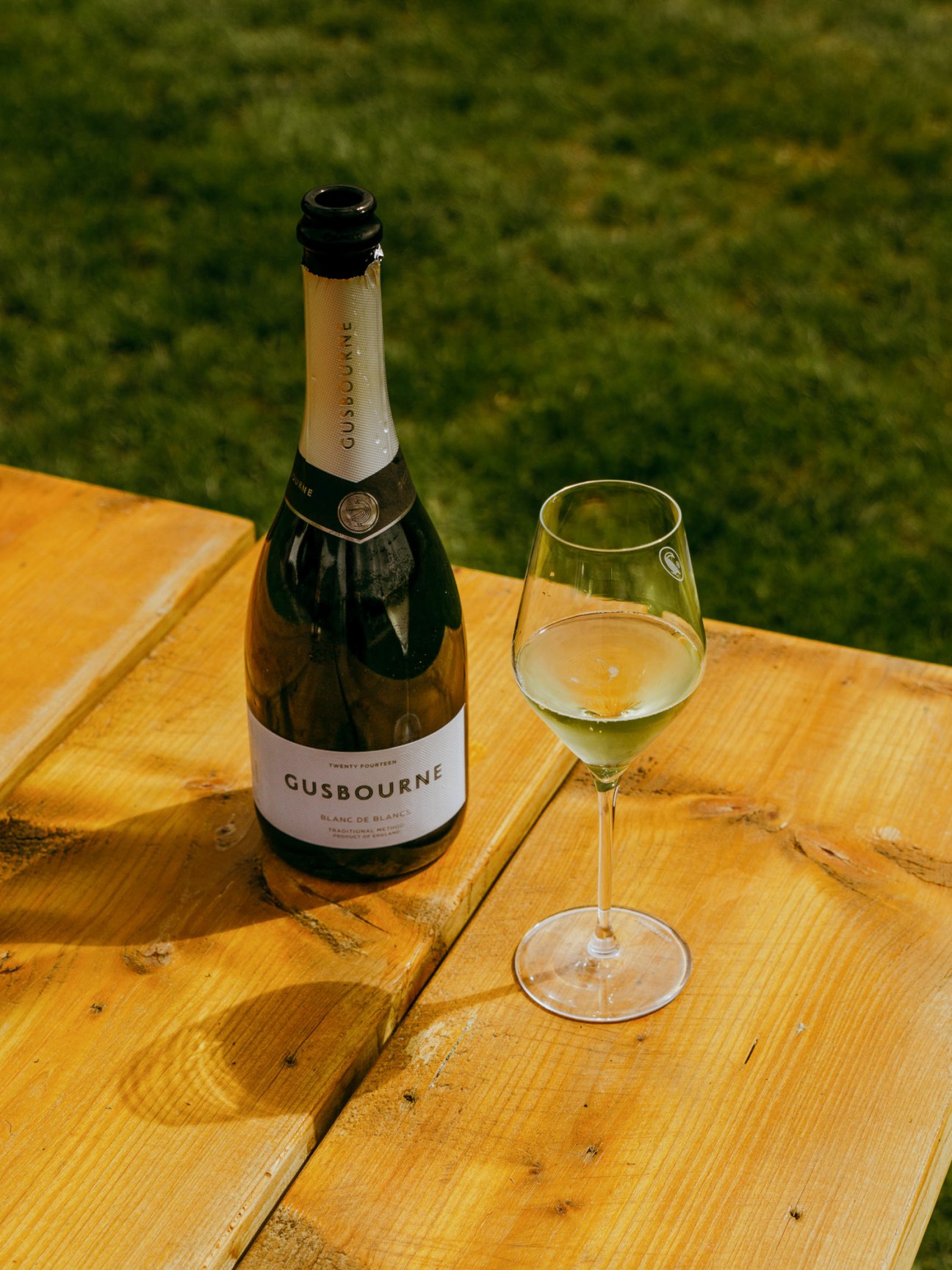 Bottle of Gusbourne sparkling wine and filled glass on wooden table outdoors