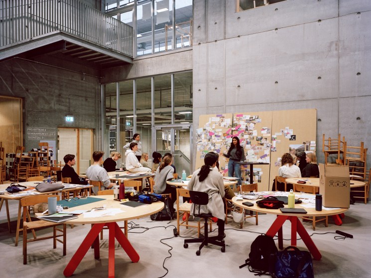Students working at wooden desks in bright concrete studio space with presentation board and stacked chairs