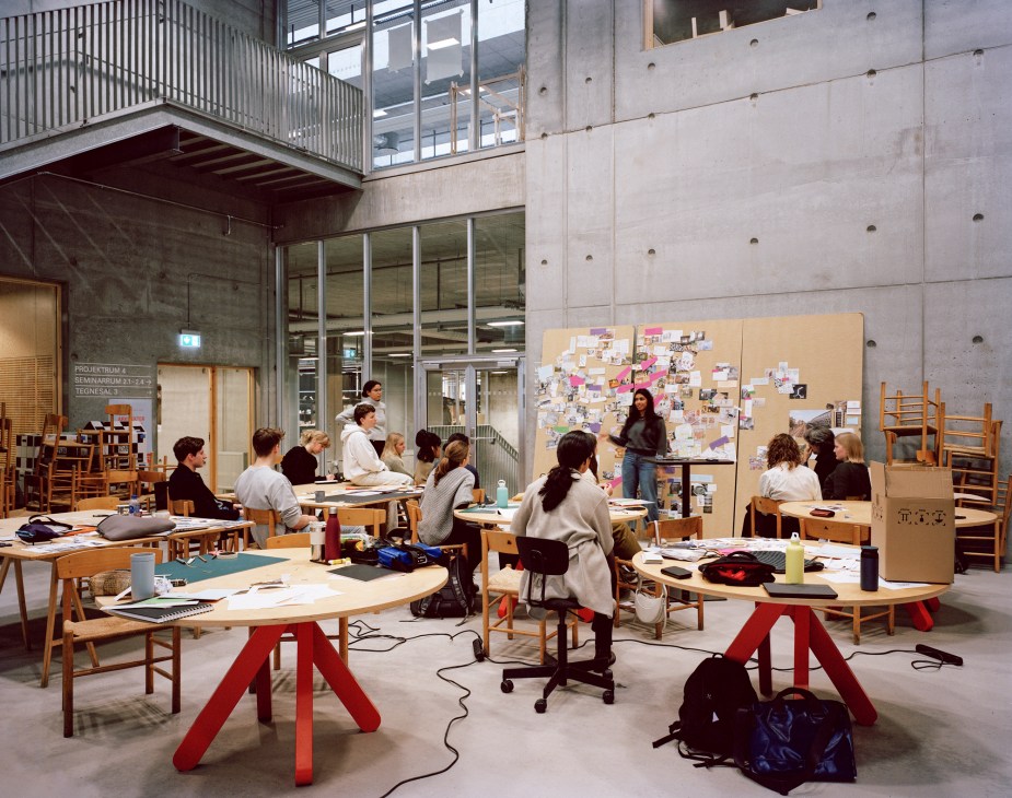 Students working at wooden desks in bright concrete studio space with presentation board and stacked chairs