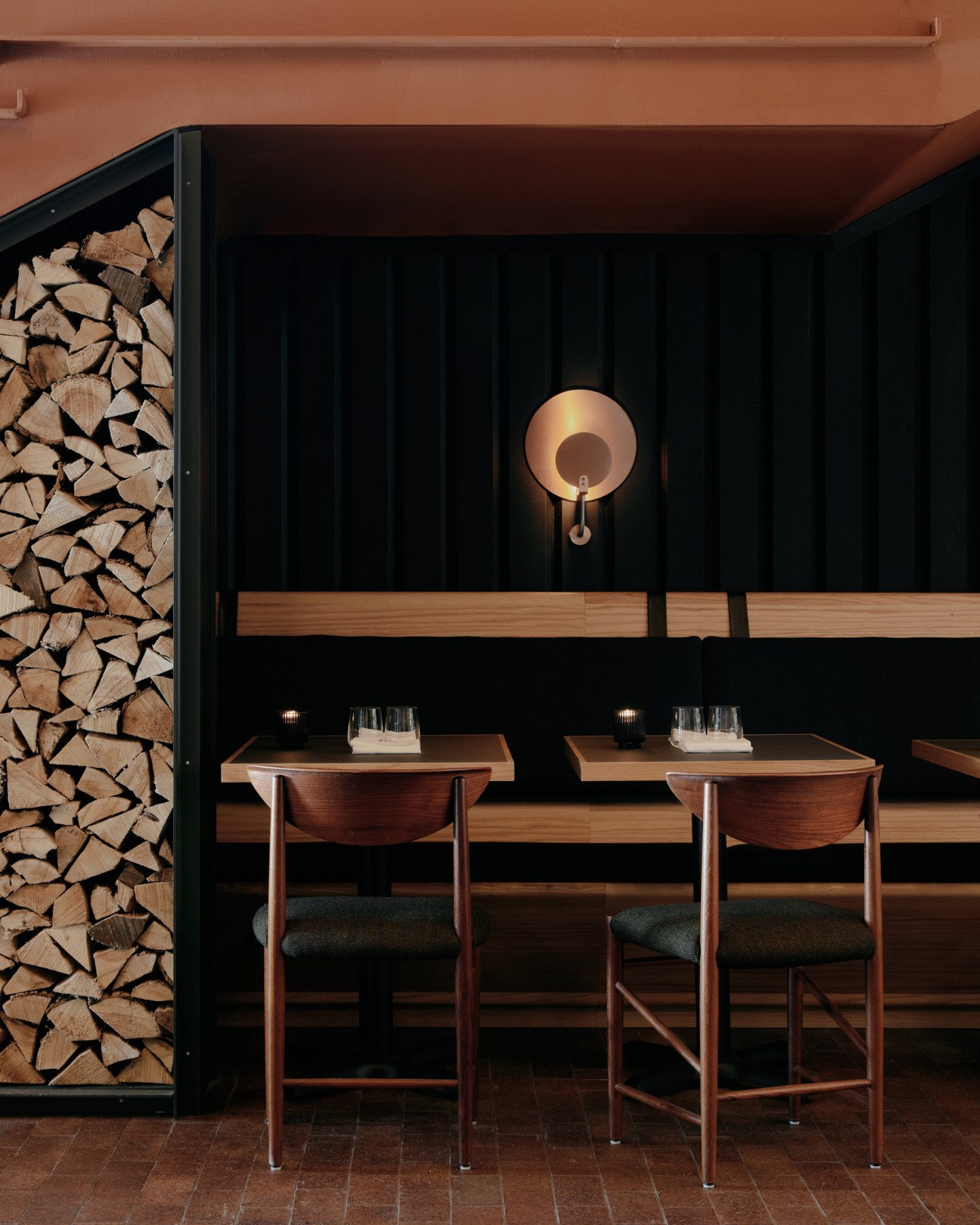 Interior dining seating at Alder restaurant with wooden tables, upholstered chairs, and stacked firewood storage