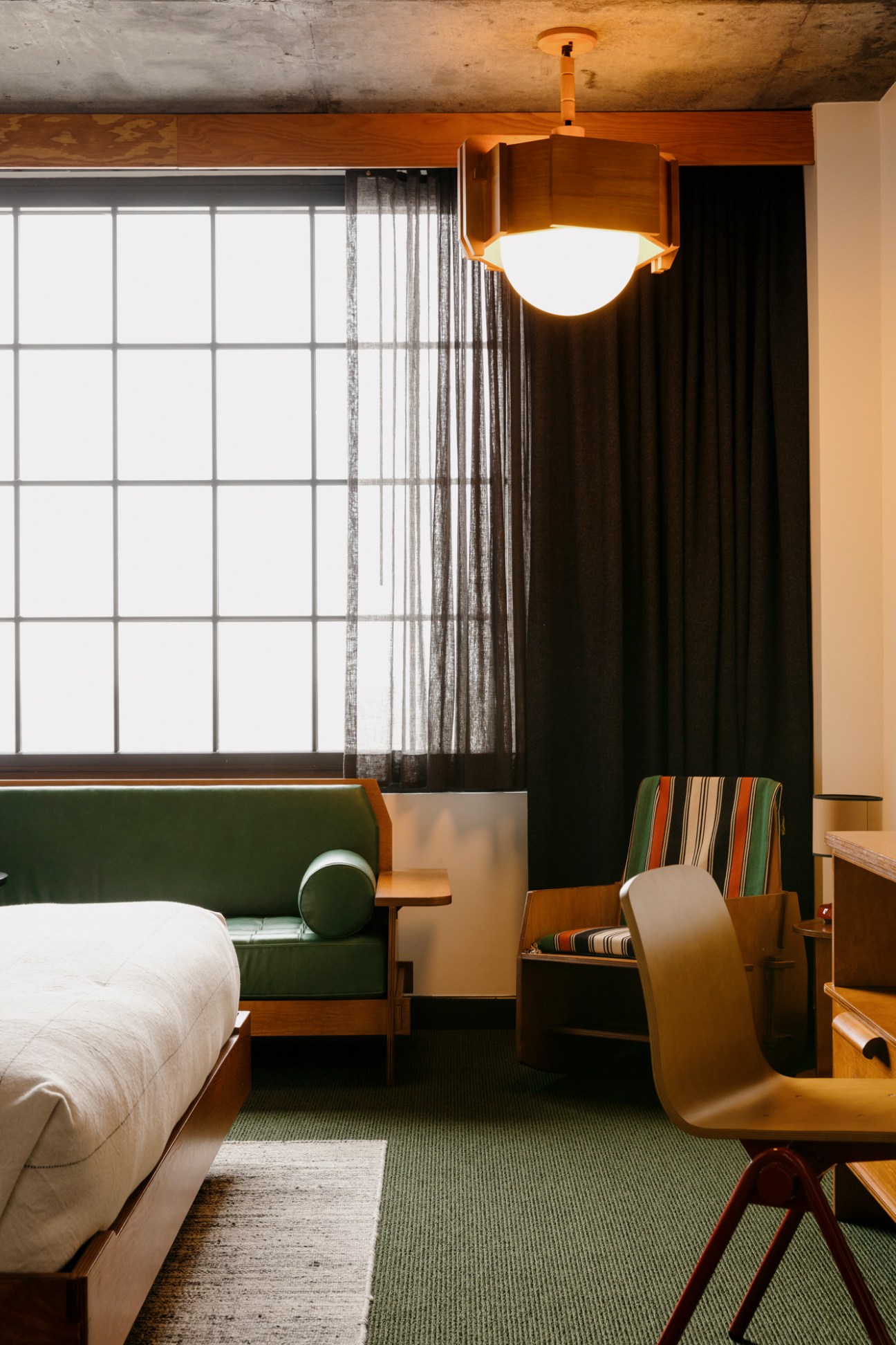 Modern hotel room with green carpet, wooden furniture, striped accent chair, and geometric pendant light