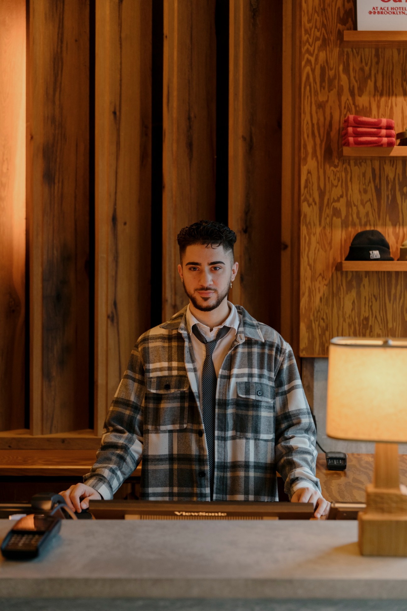 Hotel receptionist in plaid shirt stands behind wooden front desk in warmly lit lobby with wood paneling