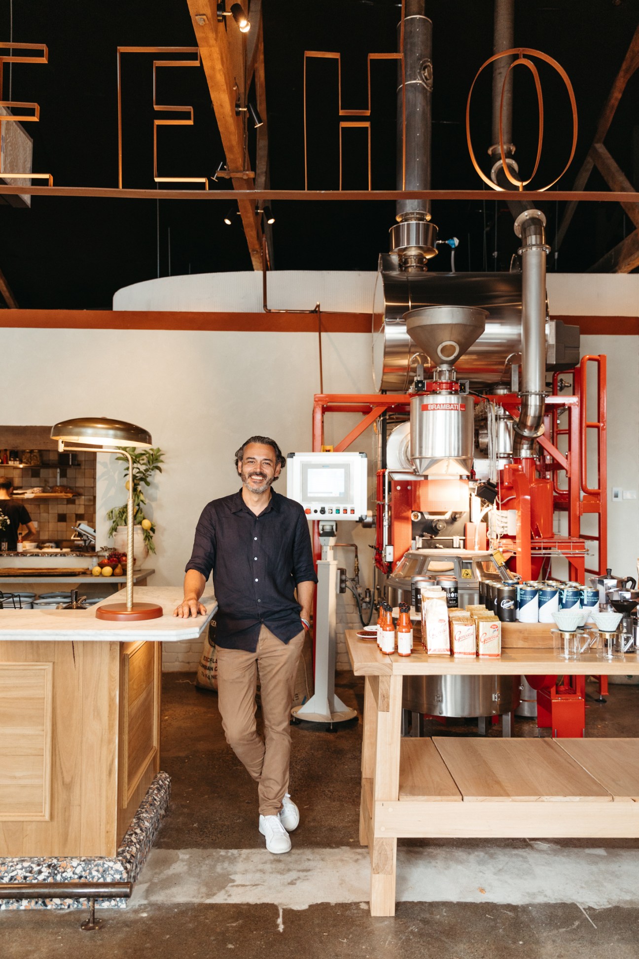 Adam Genovese standing in his coffee roastery with industrial equipment and large orange letter signage
