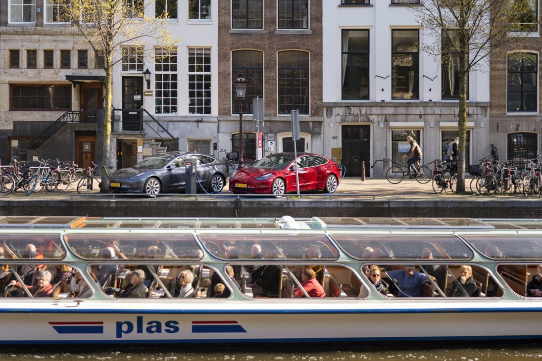 Two Tesla electric vehicles parked on an Amsterdam street lined with bicycles, with a tour boat full of passengers passing on the canal below, historic buildings in background