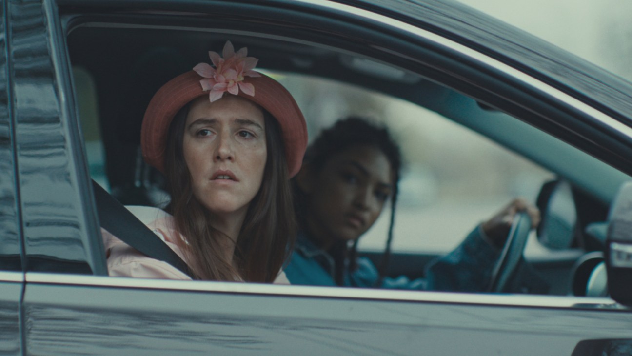 Young woman with flower-adorned hat sits in passenger seat of car looking concerned while driver is blurred