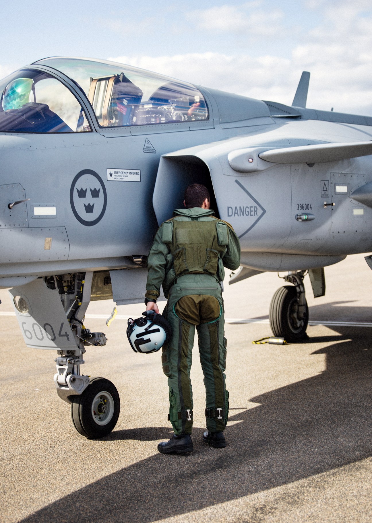 Maintenance technician in green flight suit walking toward gray Gripen E fighter jet on tarmac
