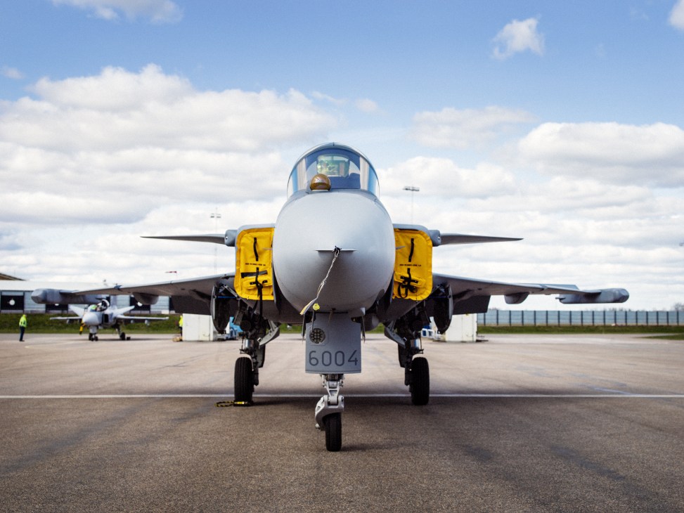Gripen E fighter jet parked on tarmac with yellow intake covers under blue sky with white clouds