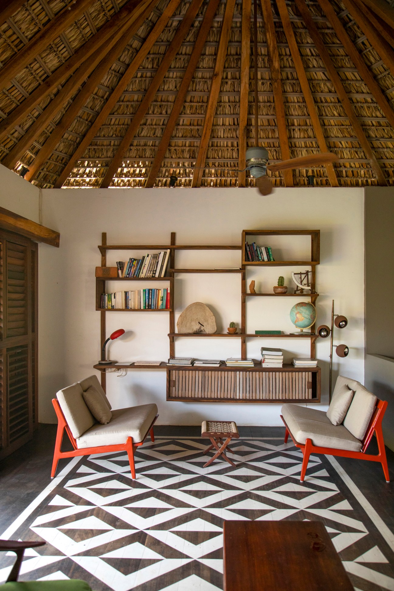 Interior with thatched ceiling, wooden shelving unit, two orange-framed chairs and geometric patterned floor
