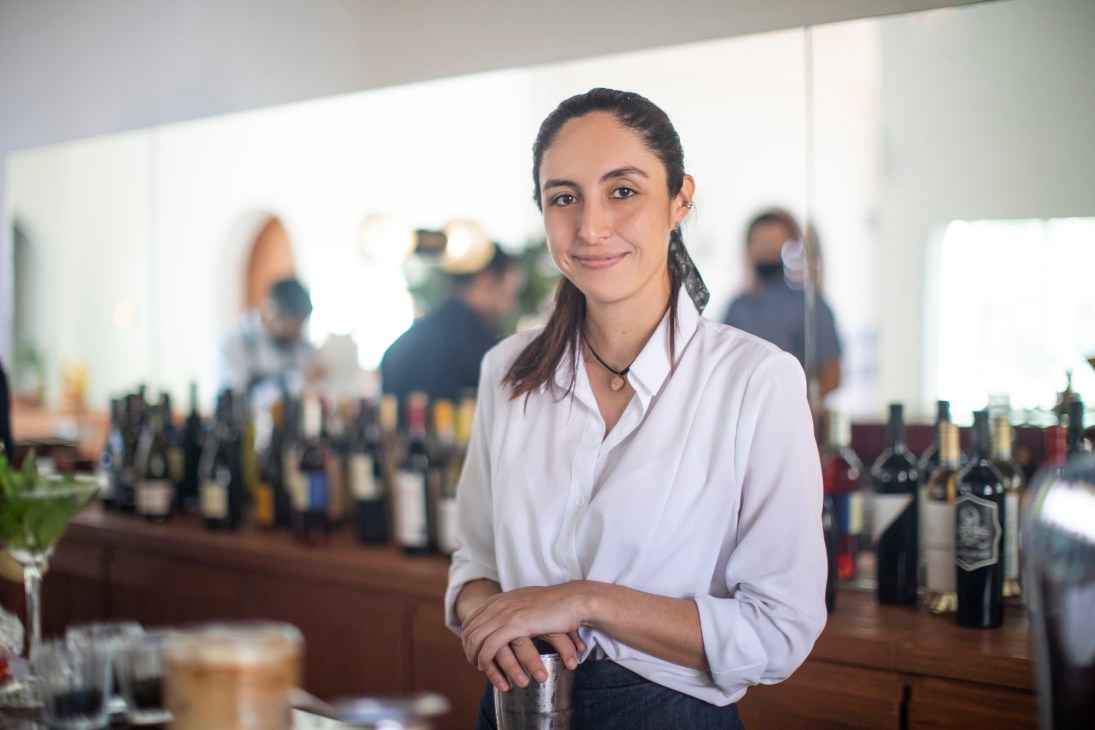 Baja Club hostess Fernanda stands at the bar in a white shirt, with wine bottles displayed behind her.