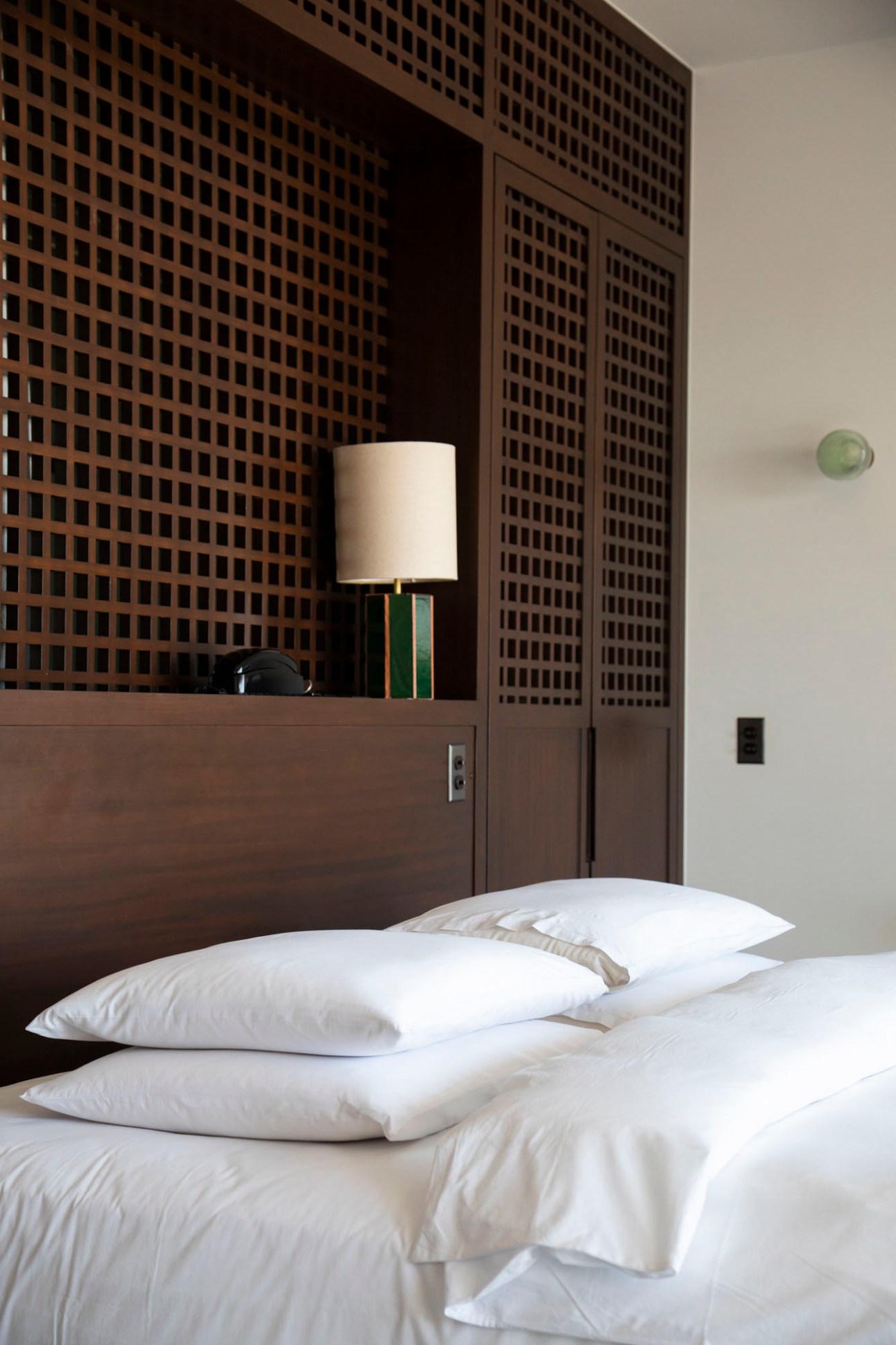 Modern hotel bedroom with white pillows on bed, wooden headboard, and latticed wall panels behind a table lamp.