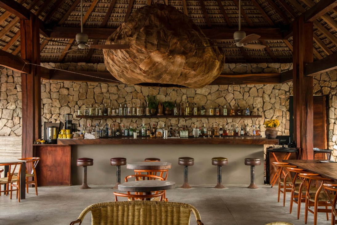 Interior of rustic bar with stone walls, thatched roof, wooden stools and bottles displayed on shelves