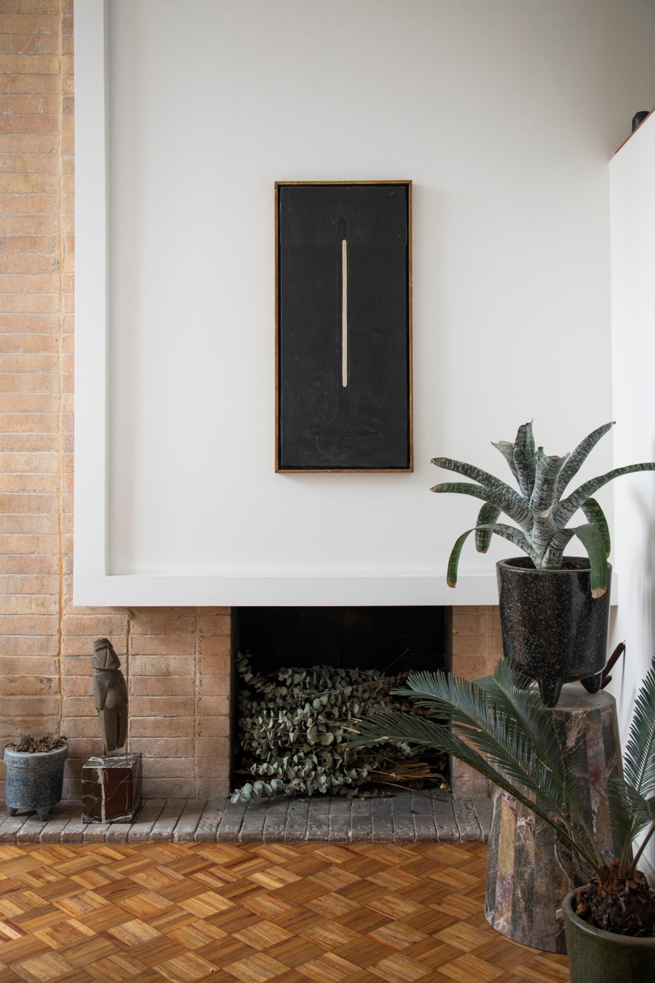 Minimalist fireplace with white mantel, framed black artwork above, plants and parquet flooring