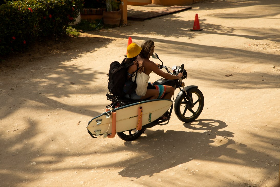 Two people riding a motorcycle with surfboard attached along a sunny road in Puerto Escondido