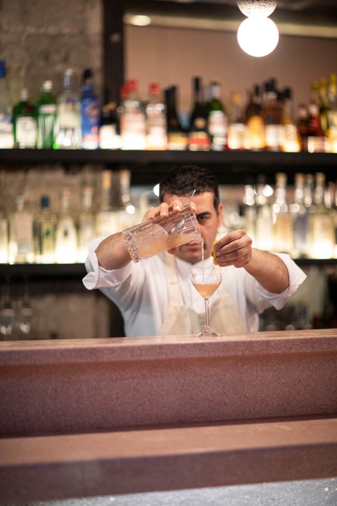 Bartender pouring a cocktail at a bar with bottles displayed on shelves behind him.