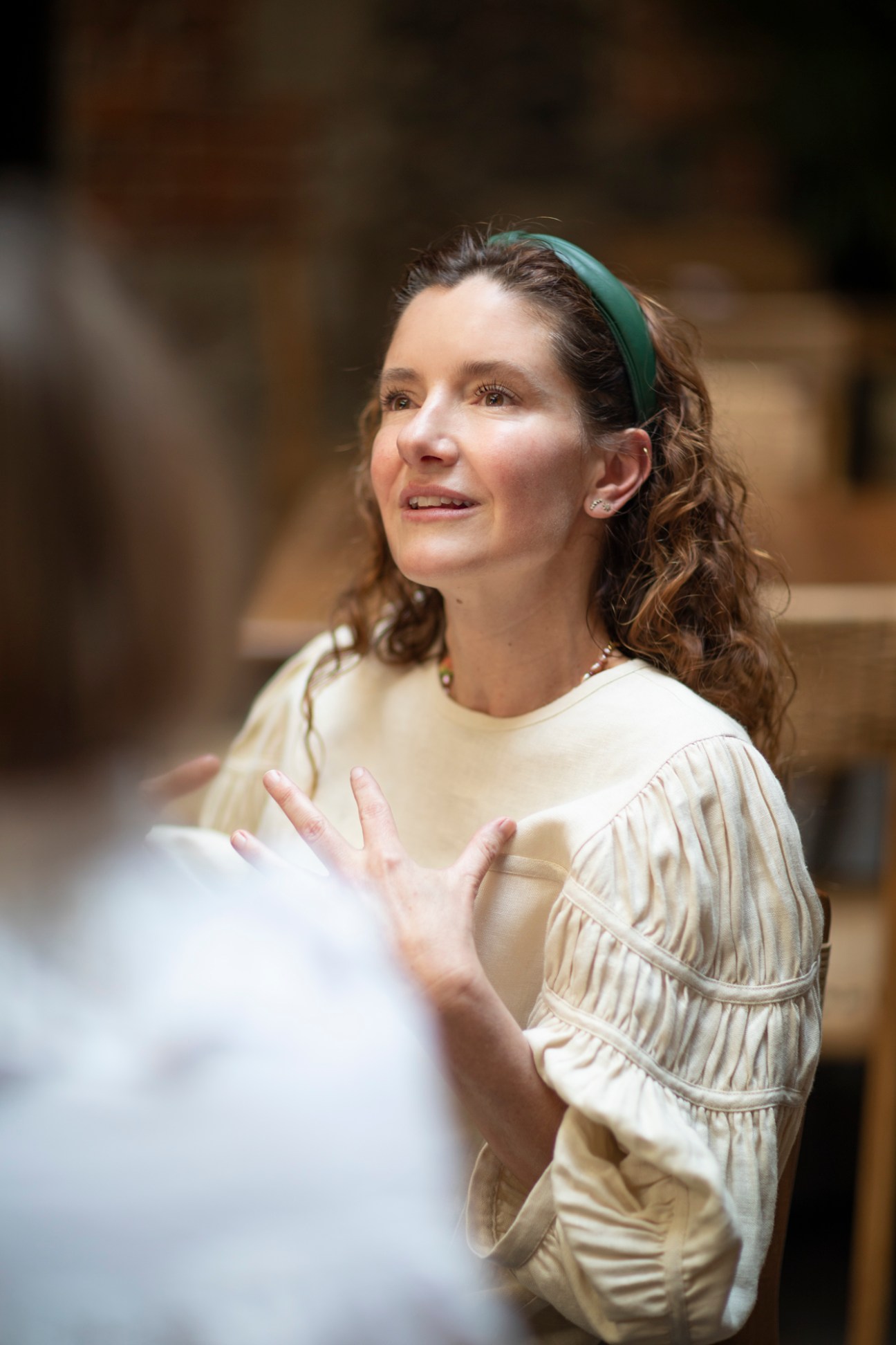 Woman in cream-colored pleated dress with green headband speaking at a table discussion.