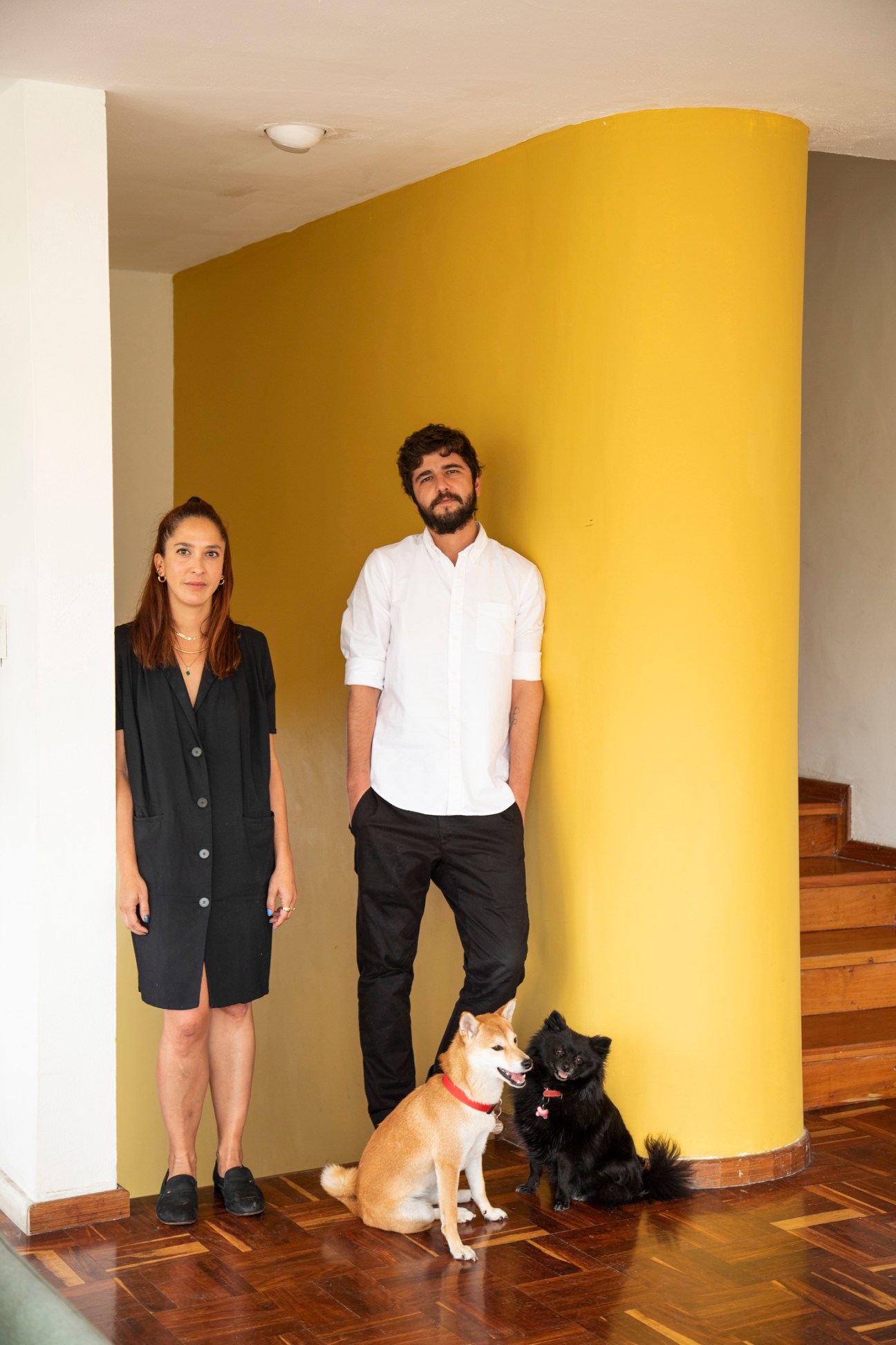 Georgina Prieto and Sebastián Vizcaino standing with two dogs in their modernist apartment with yellow curved wall