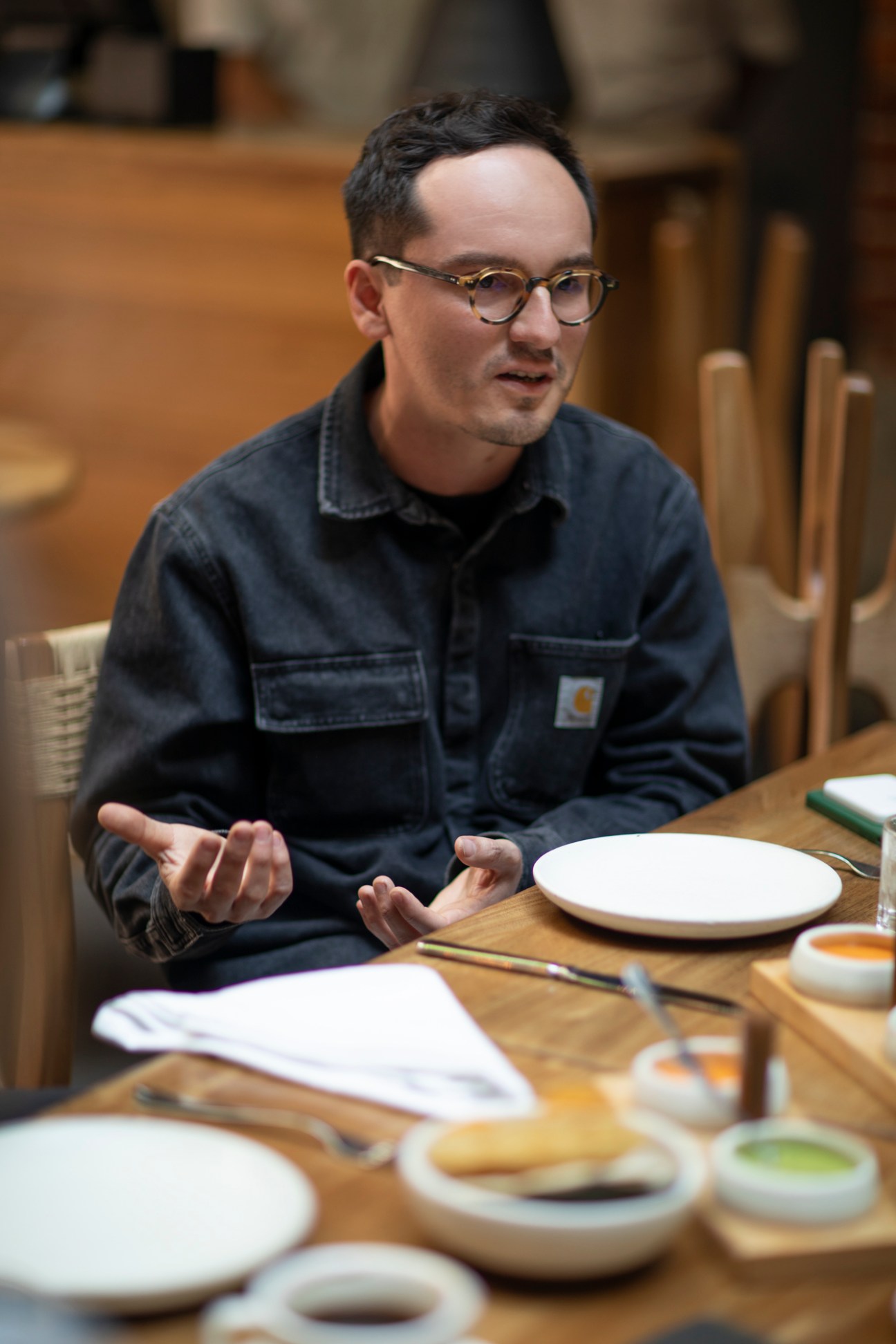 Man wearing glasses and dark denim jacket speaking at a wooden table during a round-table discussion.