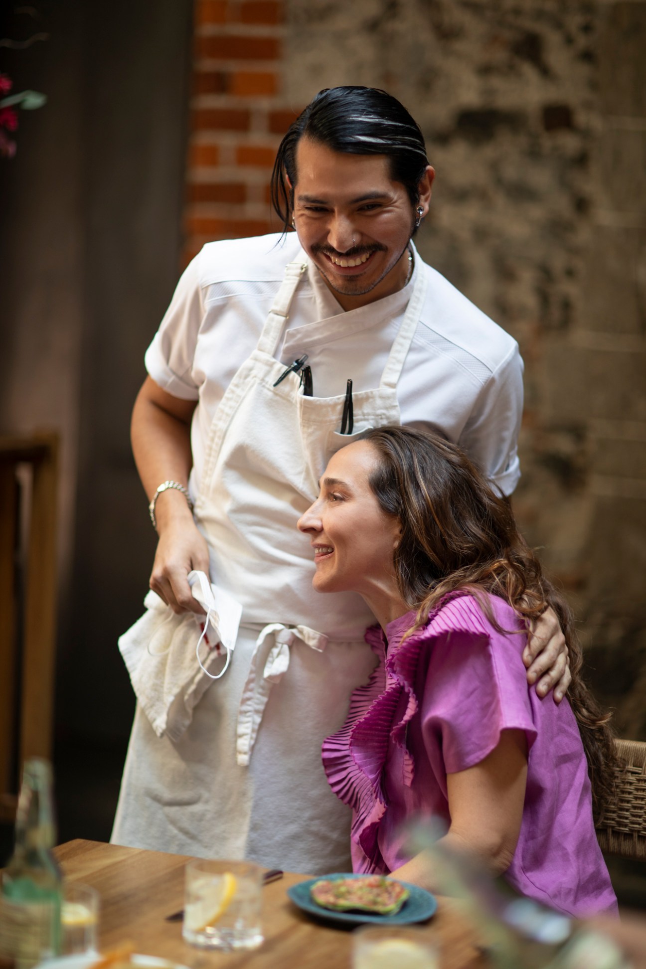 A chef in a white apron smiles while interacting with a seated female guest in a purple top at a restaurant table.