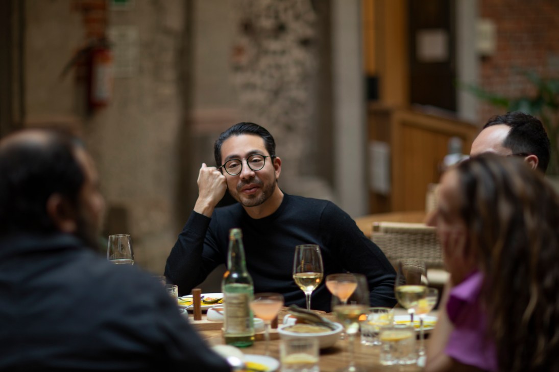 A man in glasses listens during a round-table discussion with other diners at a wooden table with wine glasses and plates.