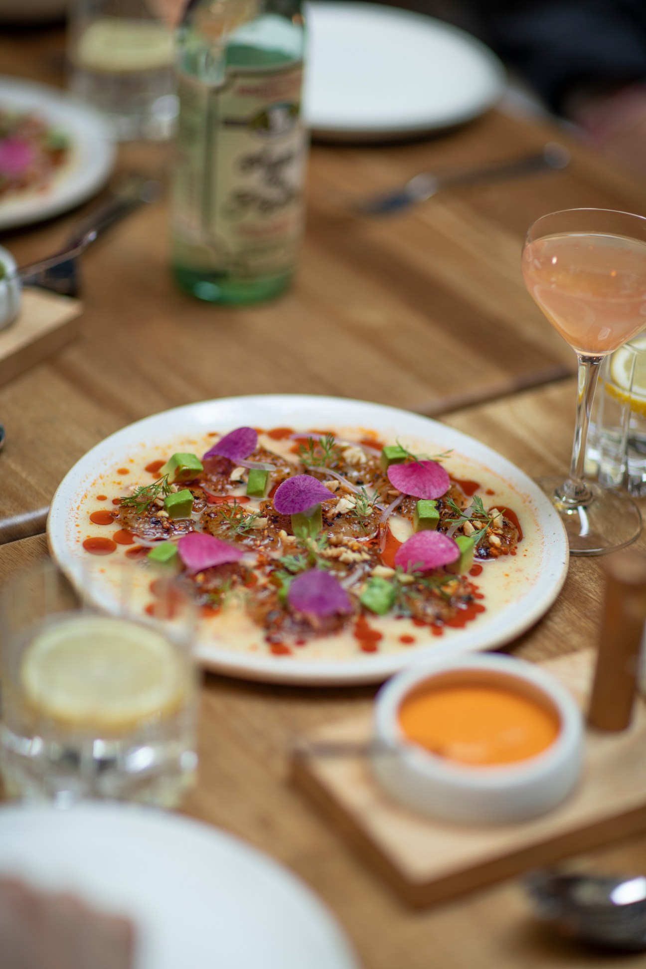 Plated dish with purple radish petals and herbs on wooden table with cocktails and condiments.