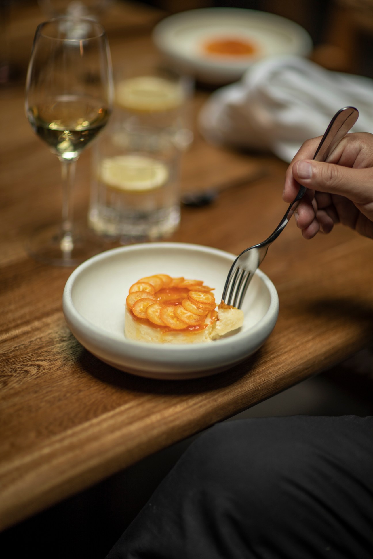 A hand holding a fork over a white plate with shrimp aguachile and sauce on a wooden table with a wine glass.