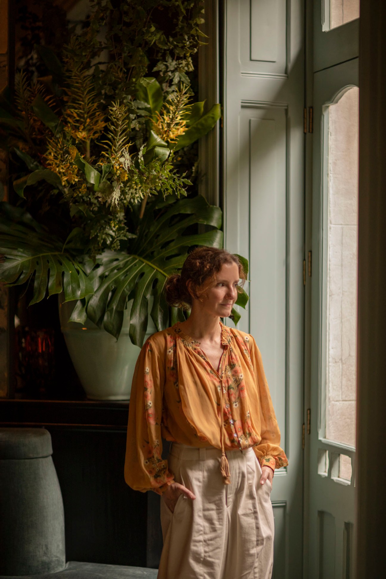 Woman in yellow floral blouse standing by window with large tropical plants in elegant green interior