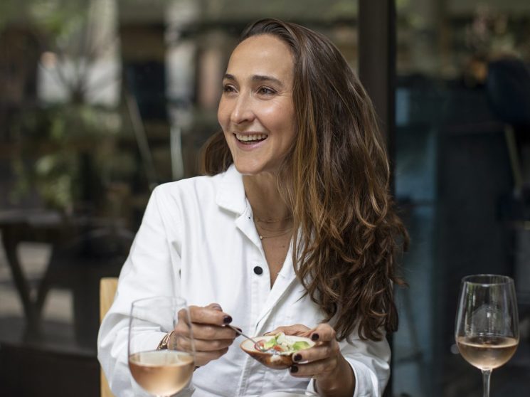 Chef Gabriela Cámara smiling while dining at a restaurant table with Mexican dishes and white wine