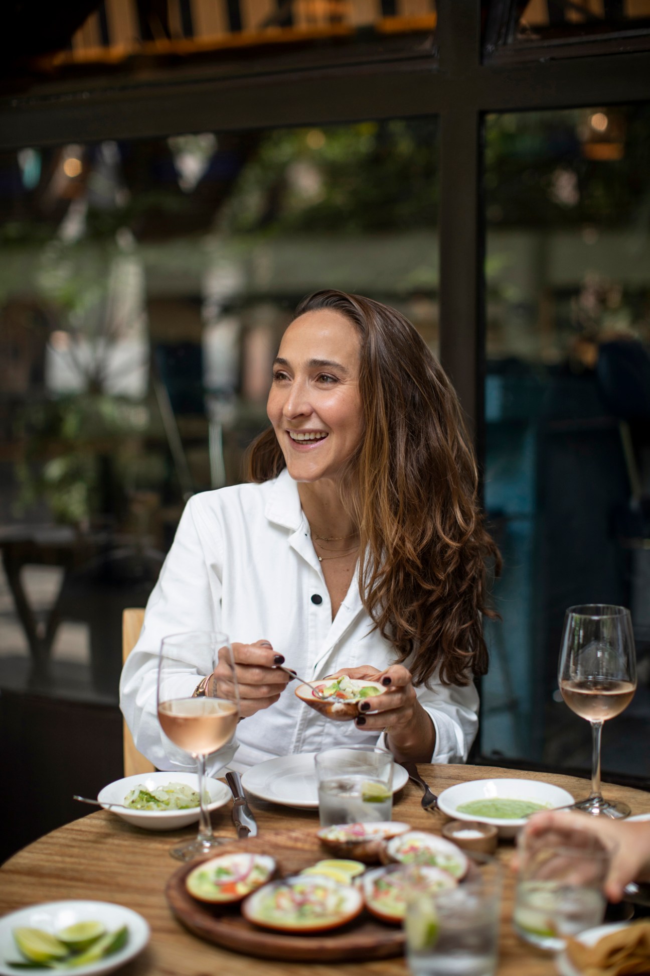 Woman in white shirt smiling while eating at restaurant table with food and wine glasses