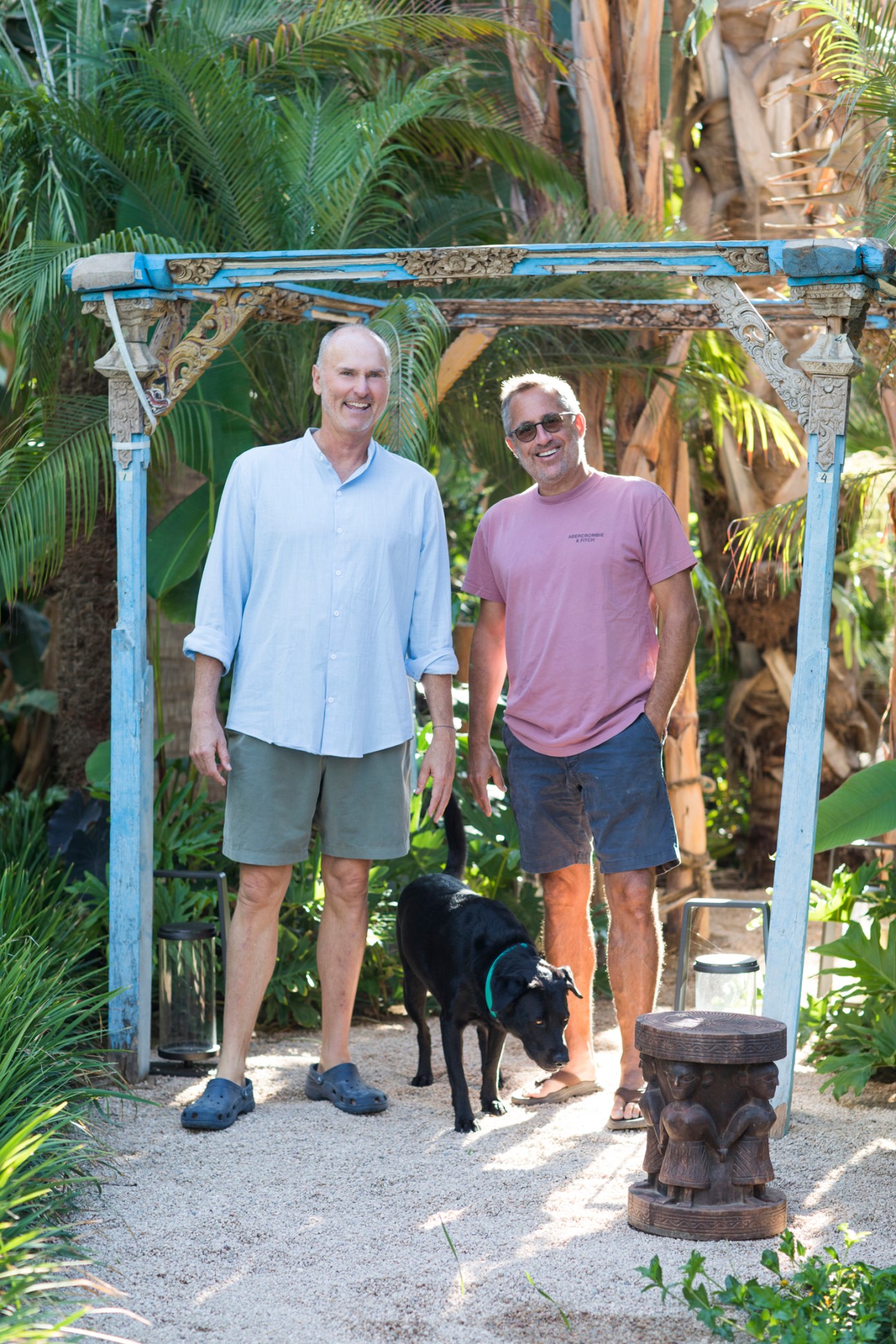 Chip Conley and interior designer Oren Bronstein standing under decorative archway with black dog in tropical setting