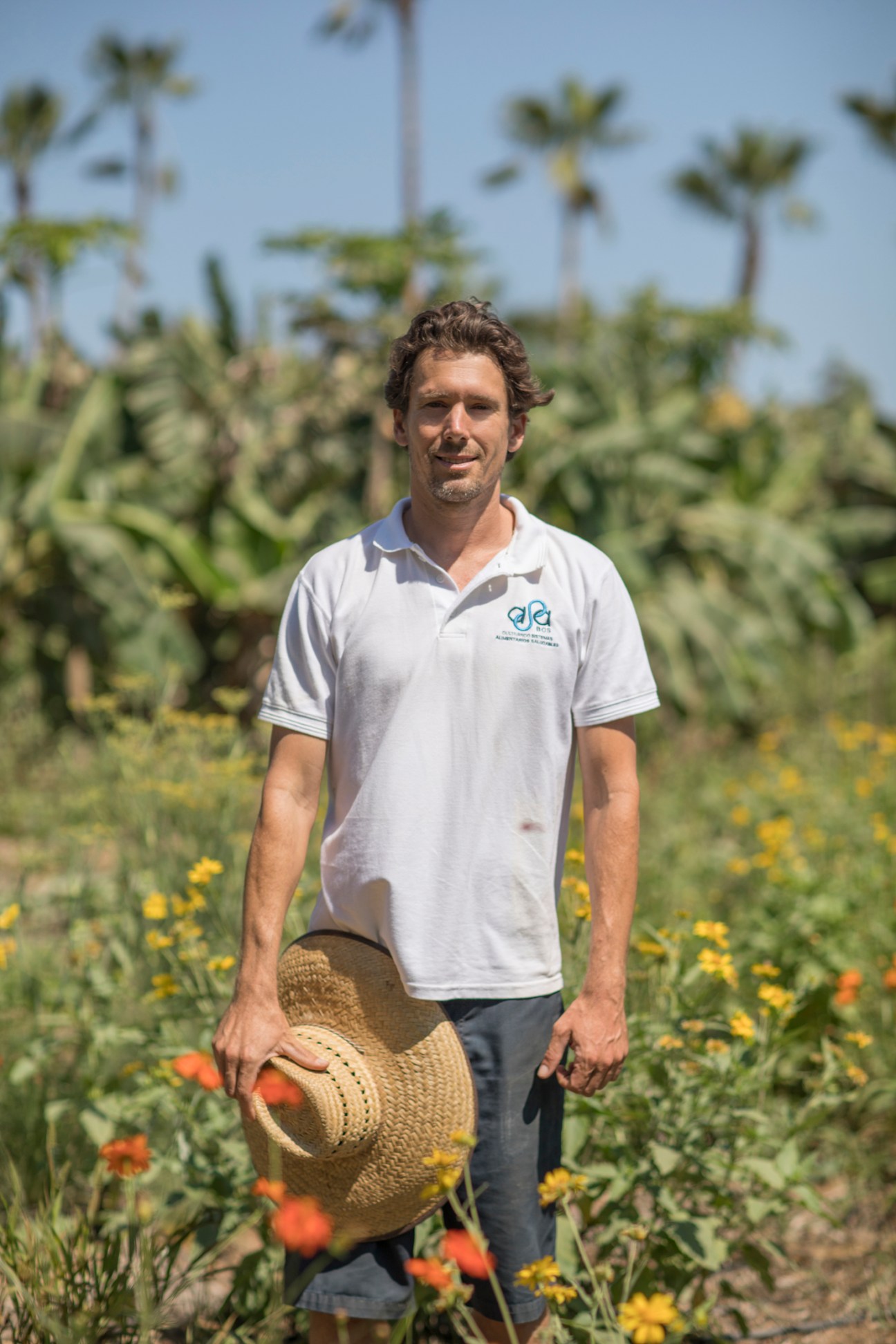 Man in white polo shirt holding straw hat stands smiling in garden with wildflowers and palm trees