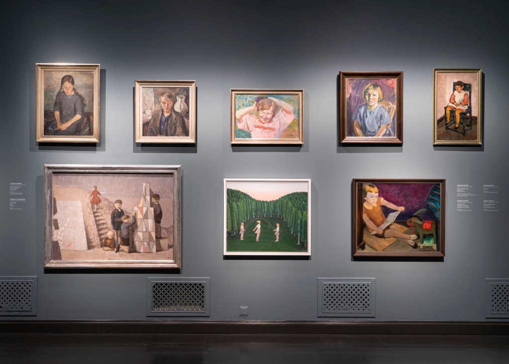 Gallery wall displaying multiple framed paintings of various sizes arranged on a gray wall with spotlighting.