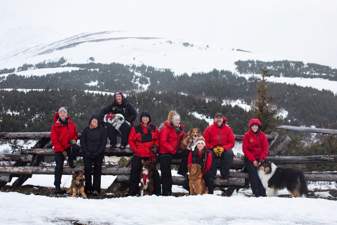 Group of Alaska Search and Rescue Dogs volunteers with their canine partners posing on snowy wooden fence