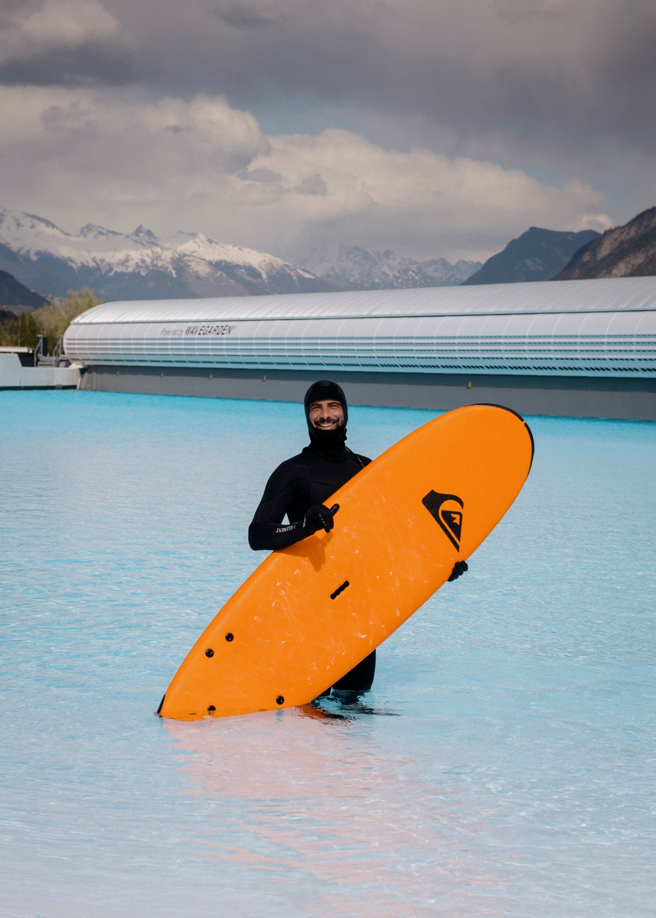 Surfer in black wetsuit holding orange surfboard in artificial wave pool with snow-capped Alpine mountains in background