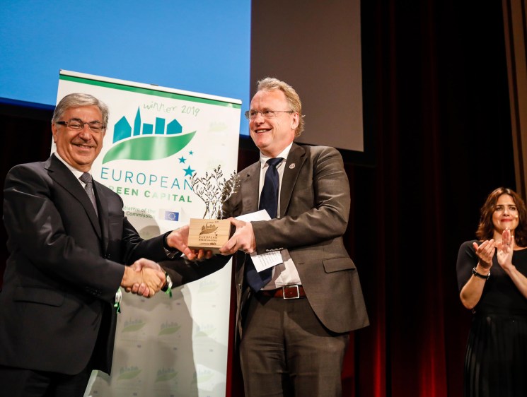 Two men shake hands on stage while exchanging an award at the European Green Capital ceremony, with a woman applauding in the background.