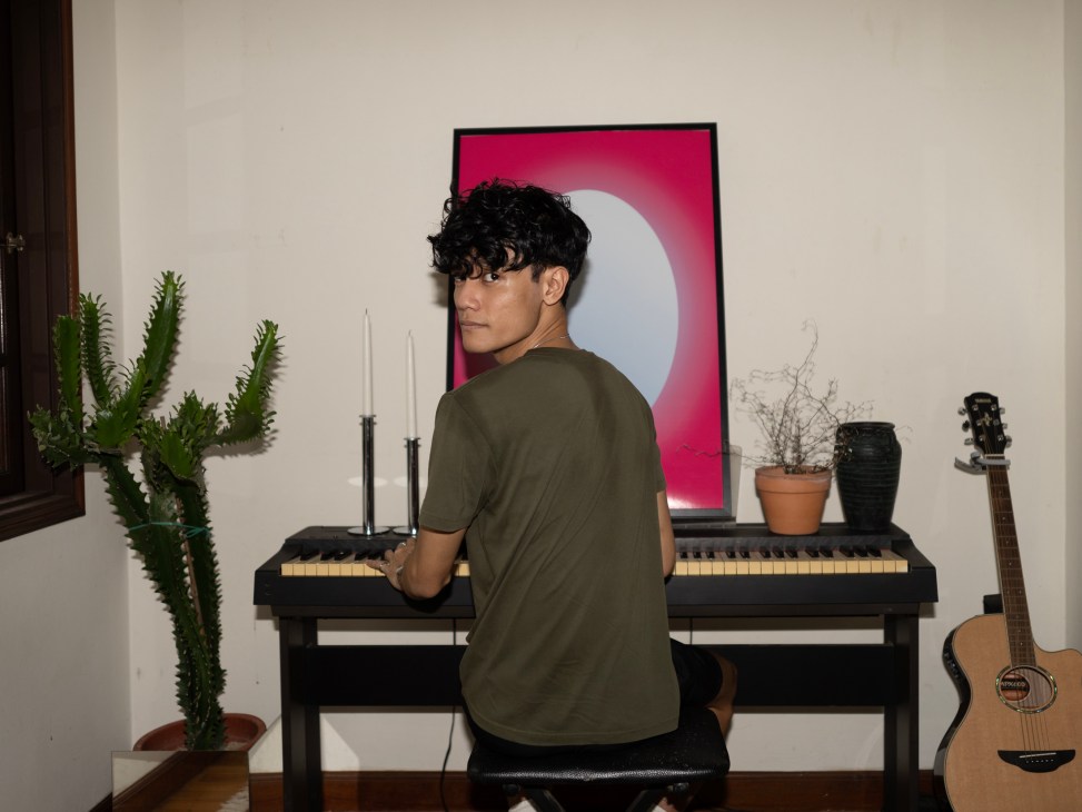 AlexTBH sits at a keyboard in his home studio, surrounded by plants, candles, and a guitar
