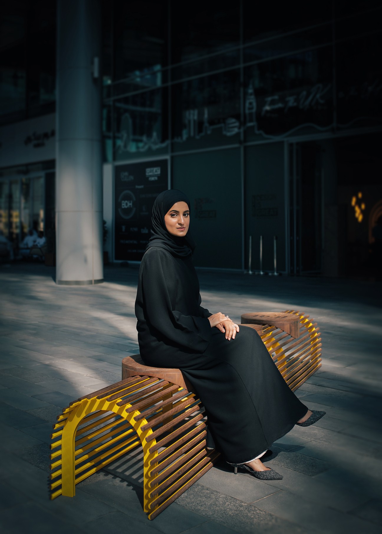 Emirati designer Aljoud Lootah sits on a wooden bench with yellow accents in an urban setting.